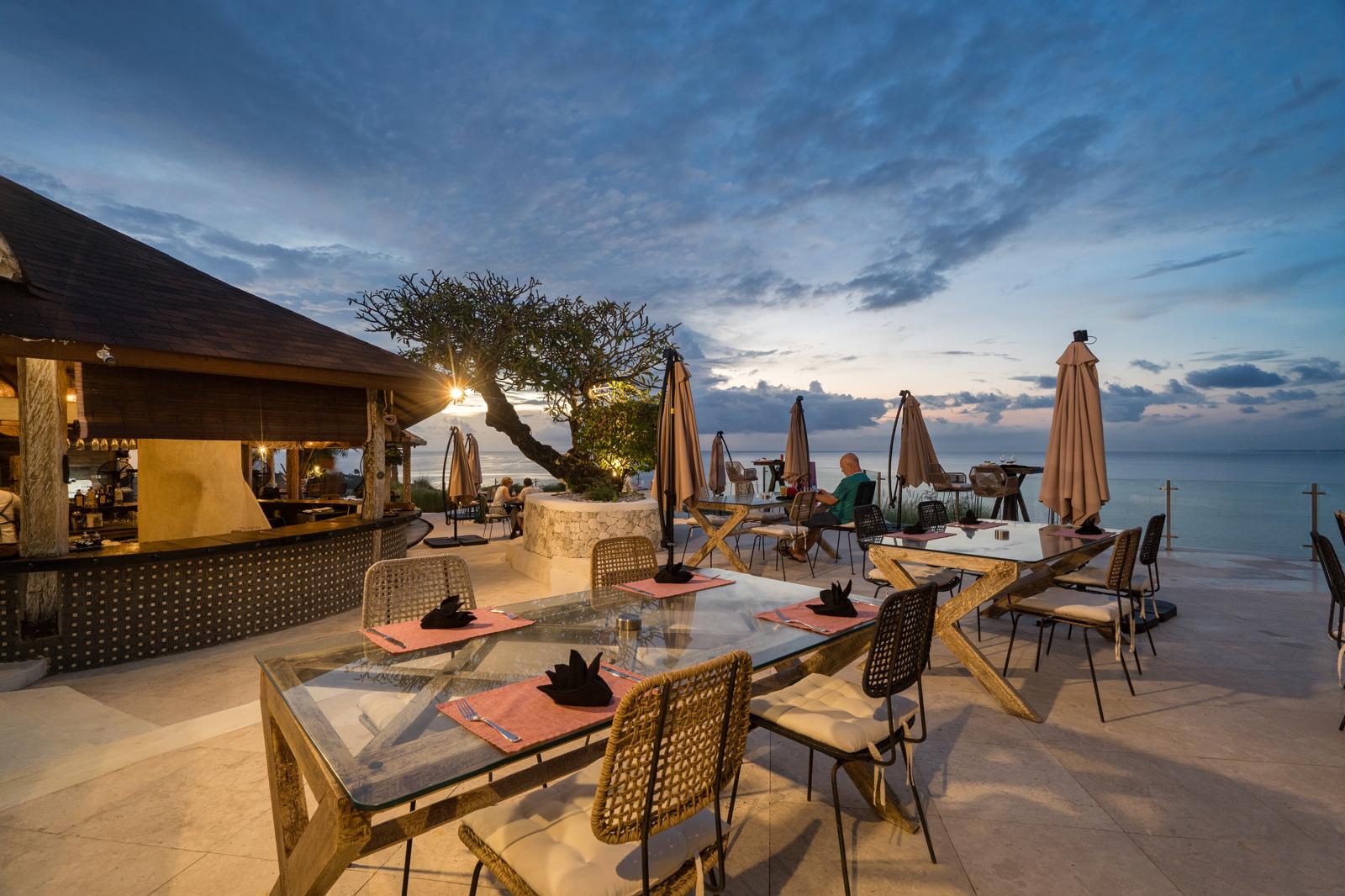 Clifftop restaurant terrace above Balangan at dusk: glass-topped tables laid for dinner with folded napkins, wicker chairs, a lit frangipani tree and the ocean behind