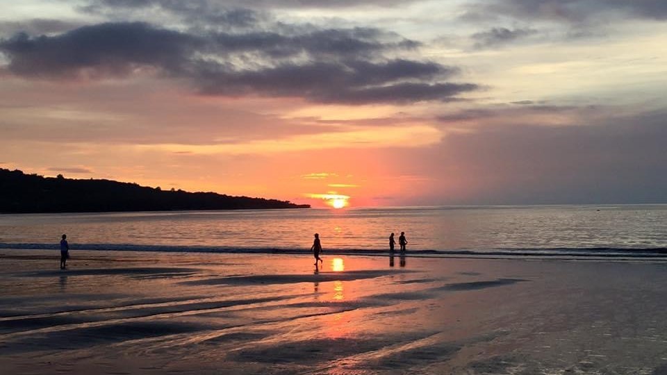 Jimbaran beach at sunset: the sun on the horizon over flat shallow water, a low wooded headland on the left, wet sand reflecting orange, a few people standing in the shallows