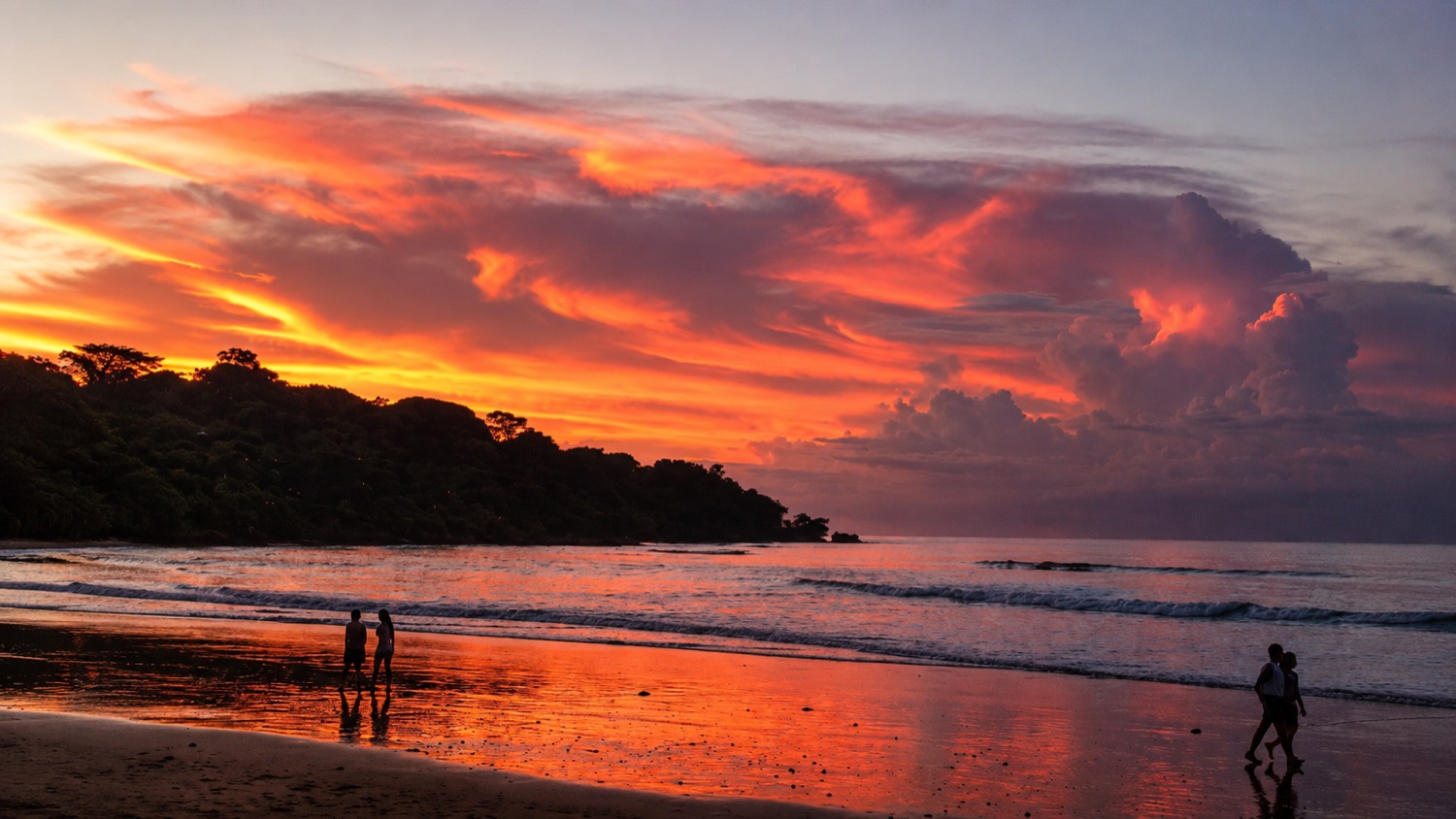 Sunset over Jimbaran beach in Bali: wet flat sand reflecting an orange sky, people walking at the water line, a wooded headland on the left