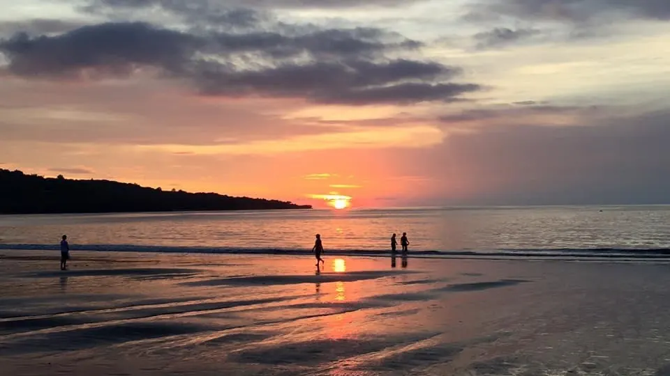 Jimbaran beach at sunset: flat shallow water, a low headland on the left, the sun on the horizon and four people standing in the shallows