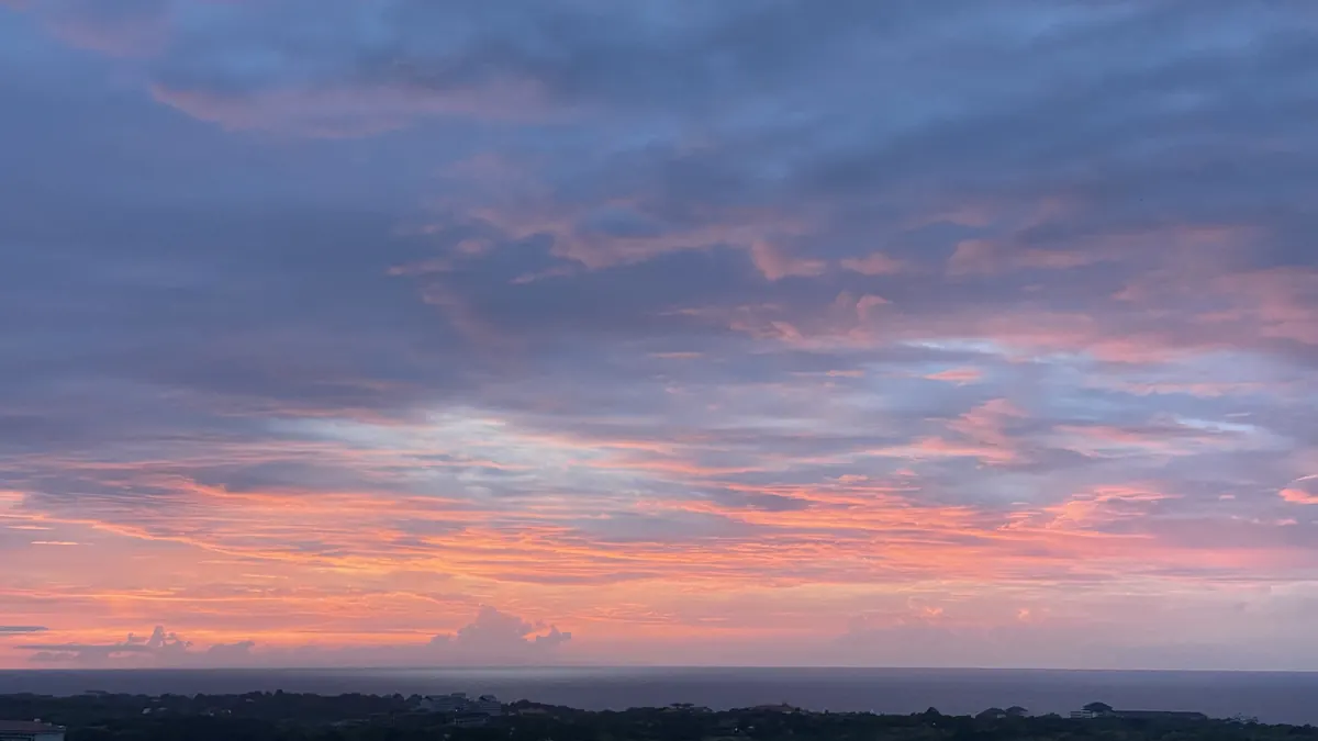 Pink and grey cloud layers over the Bukit at dusk, treeline and rooftops below, the ocean a flat line on the horizon
