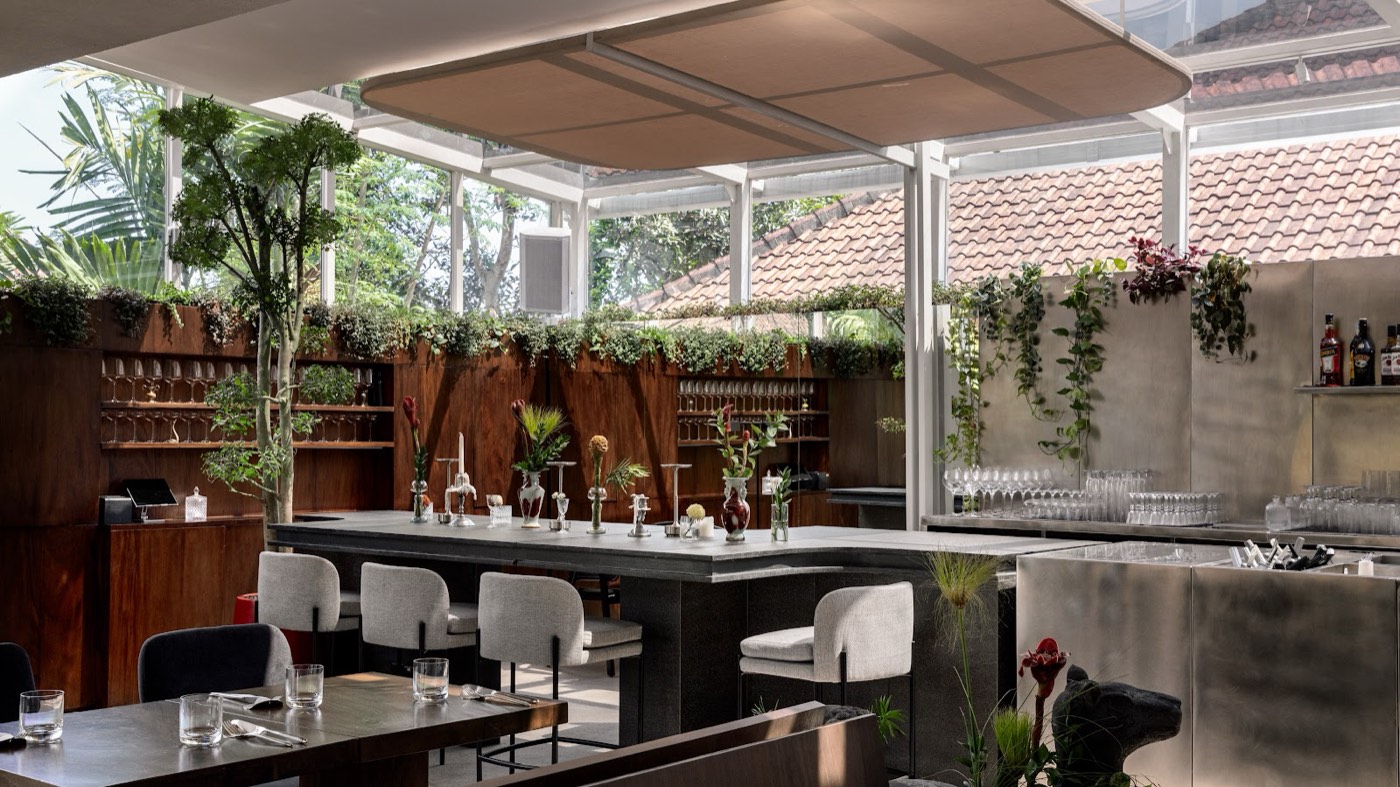 The dining room at Brie Restaurant and Cheesery in Ubud: a stone bar counter, glass walls, planting overhead and tiled roofs outside